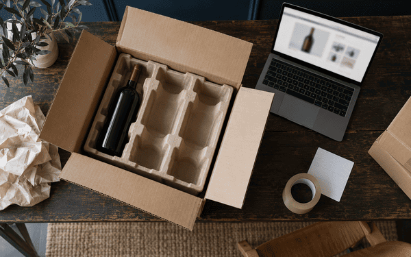 Carton d'expédition avec calage pour bouteilles et ordinateur portable ouvert sur une fiche produit, illustrant la vente de vin en ligne